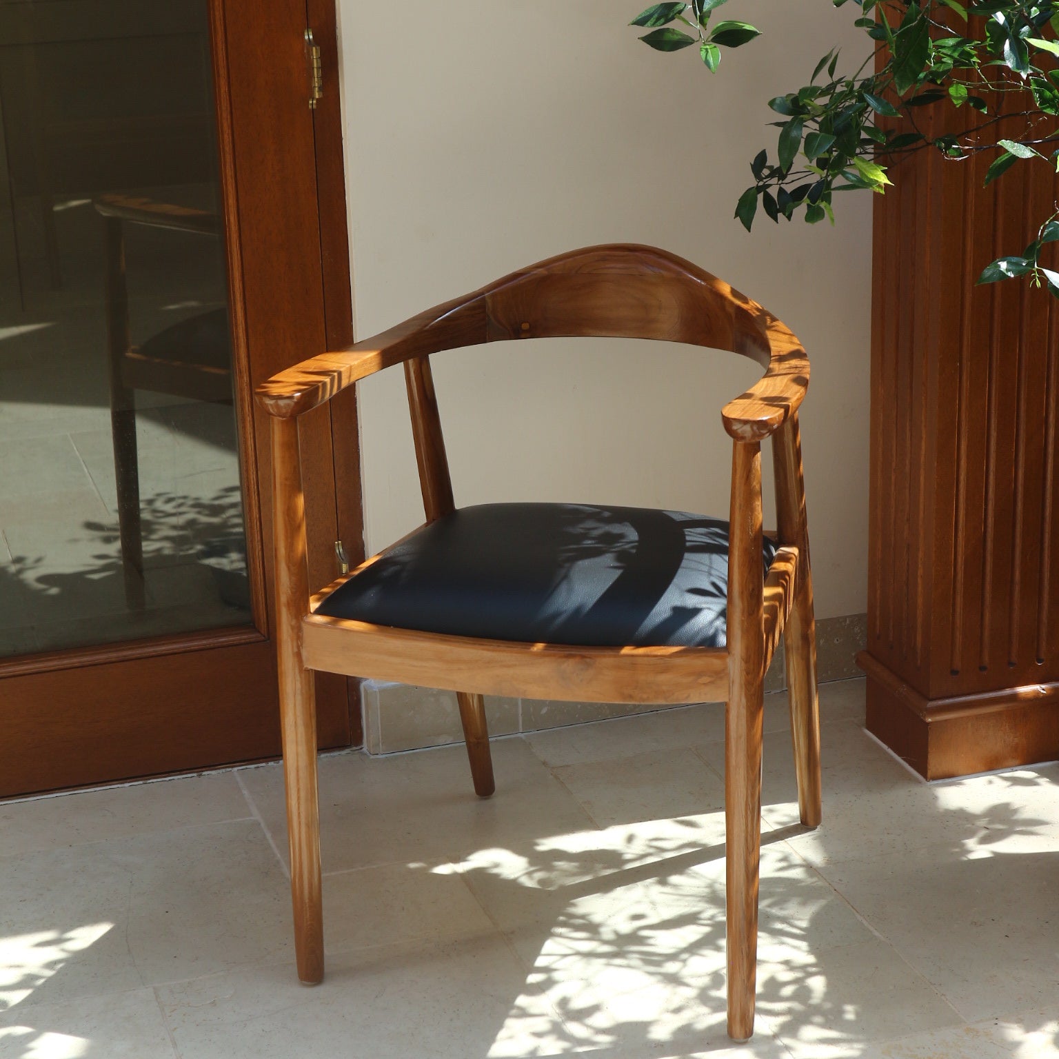 Front and side view of the Cahaya Chair, with its curved teak backrest and black upholstered seat, illuminated by soft natural light and leafy shadows
