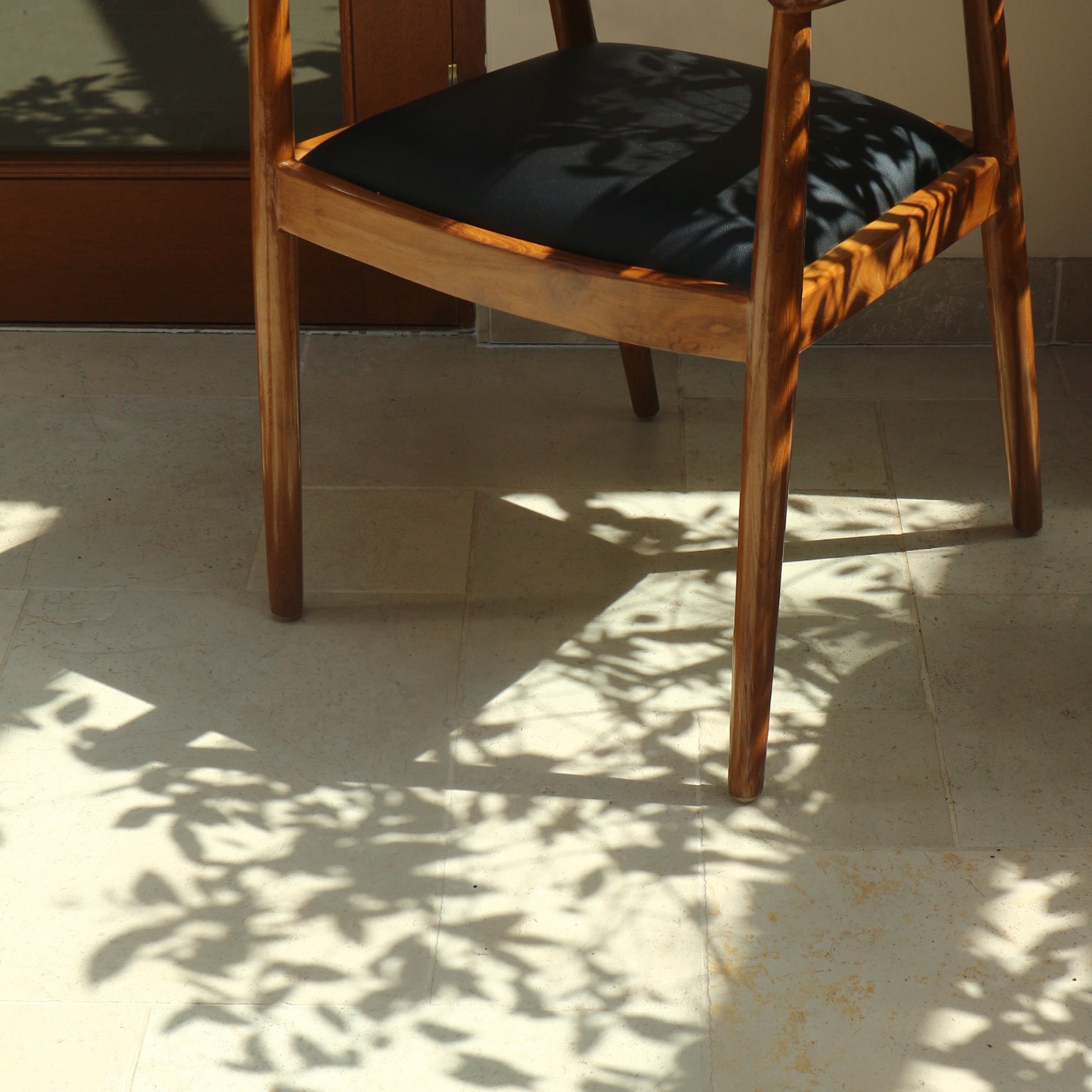 Close-up of the Cahaya Chair’s legs and black upholstered seat, with intricate shadows from nearby foliage cast across the floor.