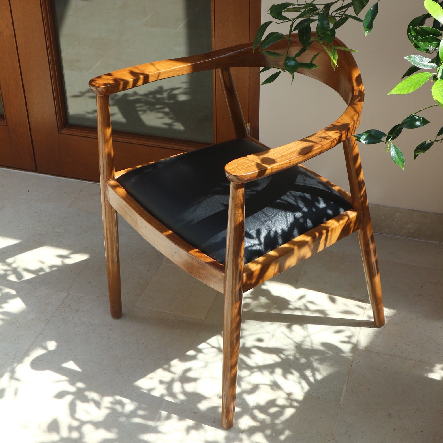 Top-angle view of the Cahaya Chair, capturing its curved teak frame and upholstered seat, with dappled sunlight and leafy shadows creating a serene atmosphere.